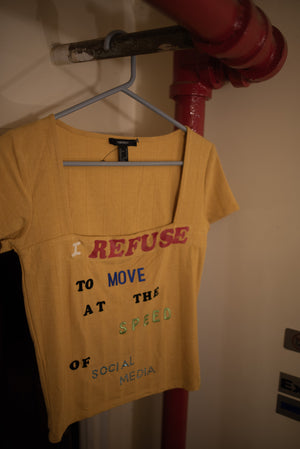 A yellow shirt with square neckline is hung on a red pipe; the shirt reads, 