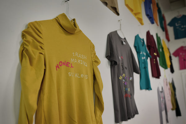 Primary colored shirts are hung on a white wall. In the foreground is a yellow turtleneck; on it reads, "Trash making honey sucklers."