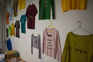 A group of shirts, in primary colors, are hung by plastic hangers that are nailed to a white wall. On each shirt is a different phrase written with felt, vinyl, and sequined iron-on letters. 